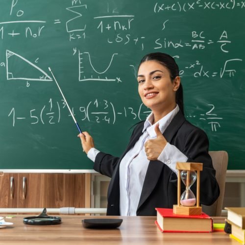 smiling young female teacher sits at table with school tools points at blackboard with pointer stick showing thumb up in classroom